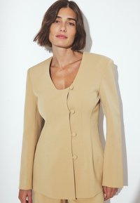 Beige tailored blazer with a v-neckline, long sleeves, and five buttons down the front. Smooth texture and structured fit highlight design.