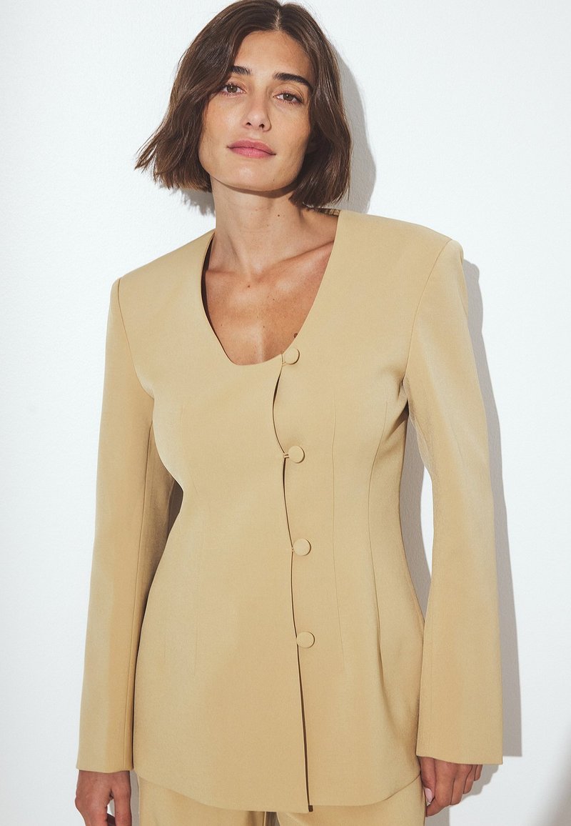 Beige tailored blazer with a v-neckline, long sleeves, and five buttons down the front. Smooth texture and structured fit highlight design.
