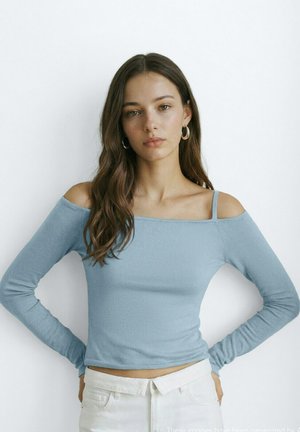 Jeune femme aux longs cheveux bruns portant un haut bleu clair à épaules dénudées et un pantalon blanc, debout les mains sur les hanches devant un fond blanc.