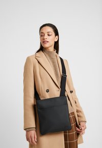 Sac besace noir en matériau texturé, avec un design plat, une bandoulière ajustable et une poche frontale, porté sur un manteau beige.