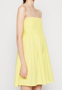 Utan axelband, smockad bodice-klänning i ljusgult bomull. A-linjeform med rynkad kjol, slät textur och längd ovanför knäna.