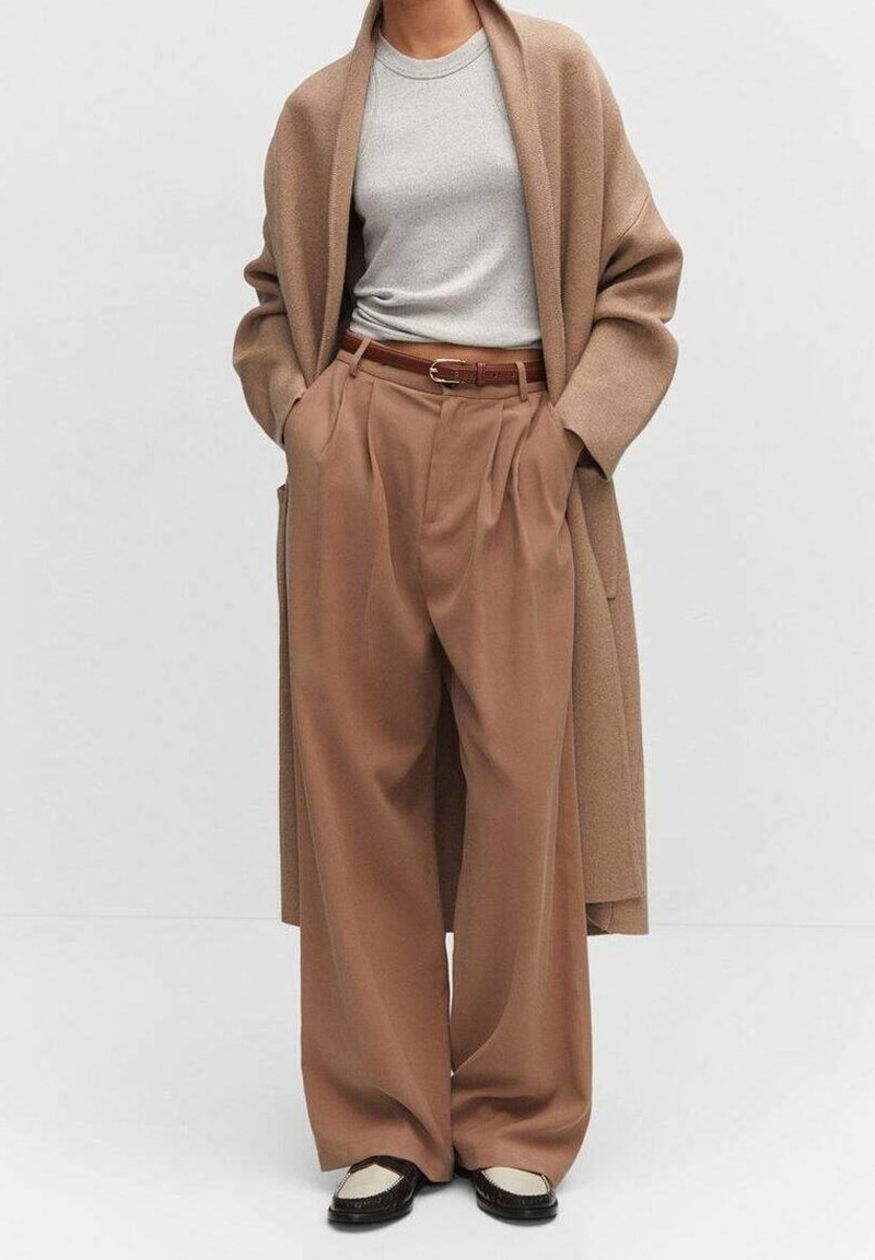 Beige wollen mantel, lichtgrijs T-shirt, hoge taille beige geplooide broek en zwarte schoenen met crèmekleurige accenten. Neutrale kleurenpalet.