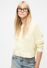 Νεαρή γυναίκα με μακριά ξανθά μαλλιά, που φορά γυαλιά με σχέδιο χελώνας, ένα ανοιχτόκίτρινο πλεκτό πουλόβερ και ανοιχτό μπλε τζιν, κοιτάζοντας στο πλάι.