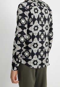 Chemise à manches longues noire avec un motif floral complexe en blanc et bleu. Matière légère avec une texture lisse et un col à revers.