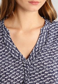 Blusa blu navy e bianca con scollatura a V e dettagli con bottoni, caratterizzata da un motivo testurizzato di forme astratte. Tessuto morbido e leggero.