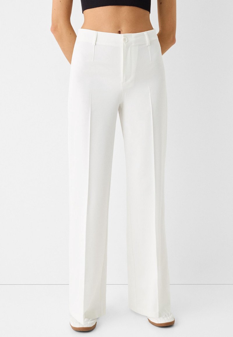 Femme portant un pantalon blanc taille haute à jambes larges avec des plis devant, associé à un top court noir et des chaussures blanches.