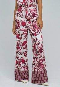 Mujer vestida con pantalones de pierna ancha y cintura alta con patrones florales y geométricos rojos, combinados con zapatos de punta blanca sobre un fondo claro.