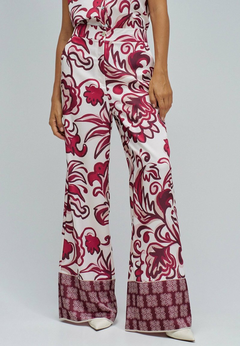 Mujer vestida con pantalones de pierna ancha y cintura alta con patrones florales y geométricos rojos, combinados con zapatos de punta blanca sobre un fondo claro.