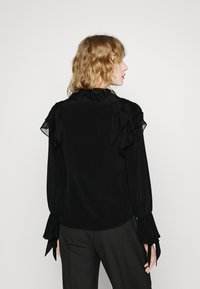 Blusa nera a manica lunga con dettagli arricciati sulle spalle e sui polsini, texture lucida e scollatura rotonda. Vista posteriore mostrata.