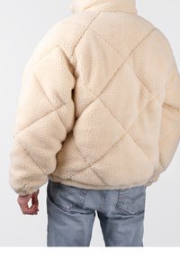 Beige quiltad fleecejacka med hög krage, texturerat tyg, puffig silhuett och elastiska ärmar, bärs över ljusa denimjeans.