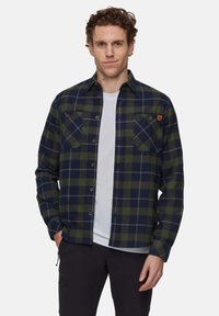 Uomo che indossa una camicia a quadri blu navy e verde sopra una maglietta bianca, con pantaloni neri, in piedi contro uno sfondo chiaro e neutro.