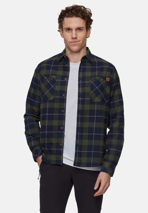 Homme portant une chemise à carreaux navy et vert, boutonnée, sur un t-shirt blanc, avec un pantalon noir, se tenant contre un fond clair.