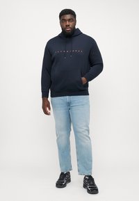 Marineblauwe hoodie met een voorzak, verstelbare trekkoordkap, bedrukte logo in oranje. Gecombineerd met lichtblauwe jeans en gestreepte zwarte sneakers.