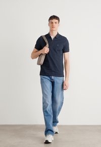 Námornícke polo tričko s krátkymi rukávmi a vreckom, skombinované s svetlo modrými džínami a bielymi teniskami. Modelka nesie šedú kabelku cez plece.