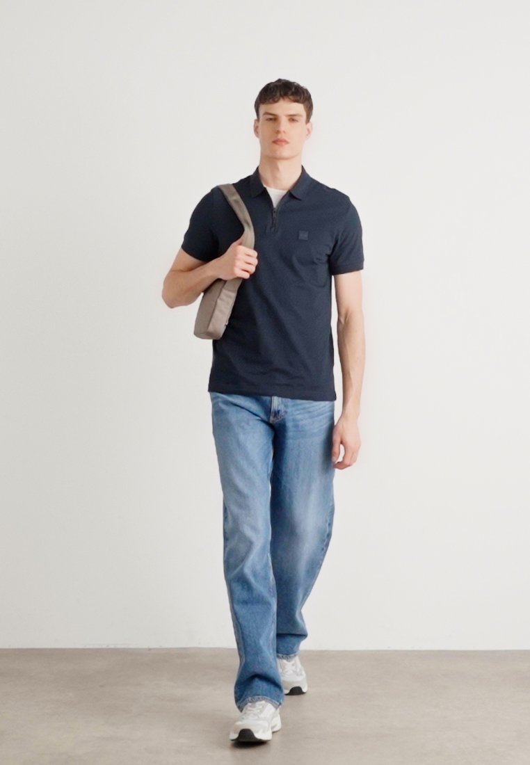 Polo marin avec manches courtes et une poche, associé à un jean bleu clair et des sneakers blanches. Le modèle porte un sac à bandoulière gris.