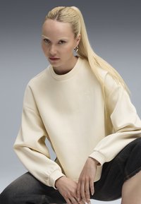 Sudadera beige claro con hombros caídos, puños acanalados y un logotipo bordado. Textura suave y corte relajado, combinada con vaqueros oscuros.
