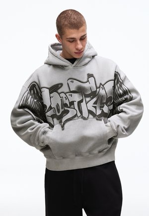 Jeune homme portant un sweat à capuche gris surdimensionné avec des textes et motifs de style graffiti noir, les mains dans la poche avant, regardant vers le bas.