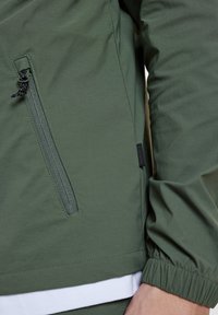 Zielona, lekka kurtka z elastycznymi mankietami, z zamykaną na zamek kieszenią i miękką teksturą. Zawiera widoczny znacznik logo.