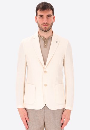 Uomo che indossa un blazer bianco sporco e testurizzato sopra una polo beige e pantaloni marrone chiaro, in piedi davanti a uno sfondo chiaro e semplice.