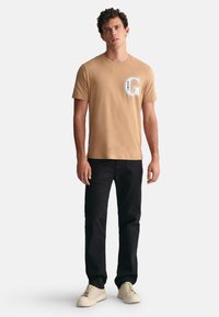 Camiseta de algodón beige con un escote redondo y un logotipo 'G' blanco impreso en el pecho, combinada con pantalones negros y zapatos de color claro.