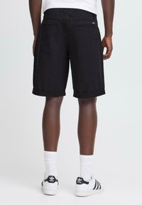 Man som bär svarta knälånga shorts, vita Adidas-sneakers med svarta ränder och vita ankelstrumpor, stående mot en enfärgad bakgrund.