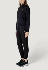 Tuta nera realizzata in tessuto morbido, con giacca zip-up a collo alto e polsini elasticizzati, abbinata a pantaloni tapered coordinati.