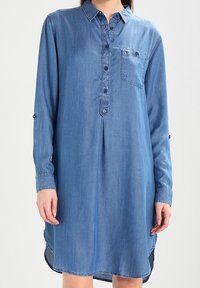 Jeanskjolklänning med långa ärmar, bröstficka, knappknäppning fram och rundad nederkant. Har en ljusblå färg och mjuk textur.