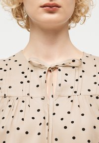 Γυναίκα με σγουρά ξανθά μαλλιά που φοράει μπεζ μπλούζα με μαύρες πουά, με δεμένο φιόγκο στο λαιμό και πιετέ λεπτομέρειες.