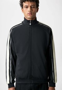 Svart zip-up träningsjacka med hög krage, med vita sidostrecker och en strukturerad tyg. Dragkedjehandtag med varumärkesdetalj.