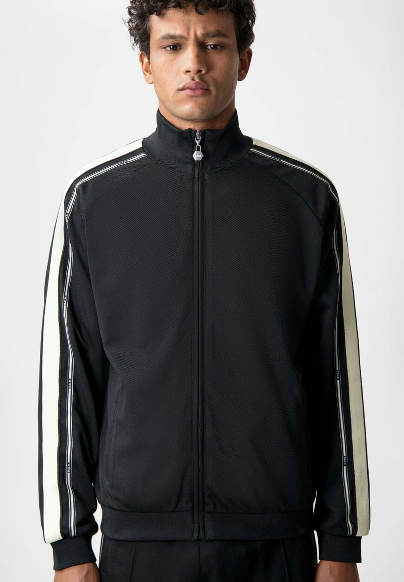 Svart zip-up träningsjacka med hög krage, med vita sidostrecker och en strukturerad tyg. Dragkedjehandtag med varumärkesdetalj.