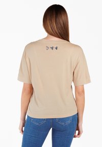 Camiseta de manga corta color beige con un corte holgado, que presenta tres pequeños corazones bordados en azul oscuro en la parte superior de la espalda.