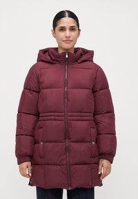 Kvinde iført en maroon pufferjakke til midt på låret med hætte, frontlynlås og trykknaplommer, stående foran en plain hvid baggrund.