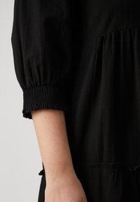 Black dress with puffed sleeves. Wrinkled texture, smocked cuff detailing, and gathered waist create a flowing silhouette.