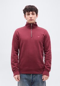 Νεαρός άνδρας φορώντας μακρυμάνικο φούτερ με φερμουάρ μέχρι το στήθος σε χρώμα βουργουνδί με μικρό λογότυπο στο στήθος, συνδυασμένο με μπλε τζιν, στεκόμενος μπροστά σε απλό φόντο.