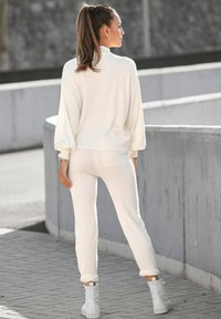 Kremsa tekstūras loungewear komplekts ar augstu kaklu un šaurām biksēm. Iezīmes mīksts audums un elastīgas piedurknes, kopā ar baltām potīšu zābakiem.