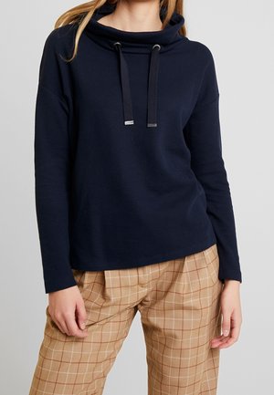 Femme portant un sweat à capuche bleu marine à manches longues avec cordons et un pantalon beige à carreaux sur un fond uni.