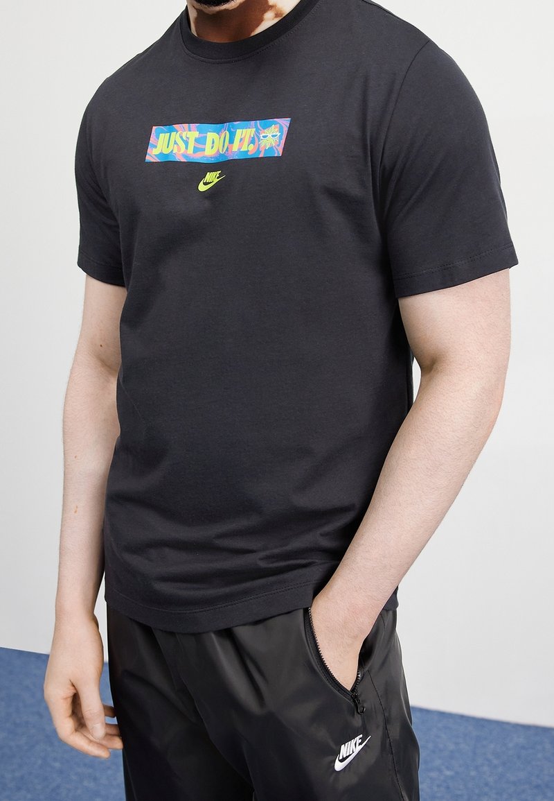 Musta puuvillane T-särk, millel on ees värviline "Just Do It" graafika ja Nike'i swooshi logo. Ümmargune kaelus ja lühikesed varrukad.