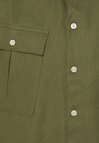 Olivgrünes Hemd aus strukturiertem Stoff, mit einer aufgesetzten Tasche und weißen Kunststoffknöpfen vorne zur Verschluss.