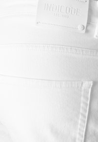 Gros plan sur la poche arrière d'un jean en denim blanc avec un patch en cuir blanc portant la marque indiquée "INDICODE EST. 1993."