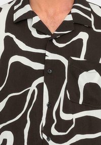 Camisa de manga corta con una base marrón oscuro y un patrón abstracto en blanco con formas onduladas. Presenta un cuello, cierre de botones y un bolsillo en el pecho.