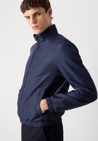 Navy blue bomber jacket made of smooth, lightweight material. Features a high collar, zip closure, and elastic cuffs. Casual design with side pockets.
