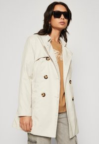 Trench coat bej cu un design ajustat, cu două rânduri de nasturi mari și o textură netedă. Purtați-l peste un top maro deschis și pantaloni gri.