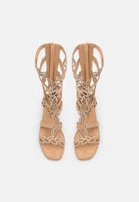 Beige gladiator-sandaalit, joissa on monimutkaisilla lehdenkaltaisilla koristeilla, nauhakerroksella, avovaosalla ja tasaisella kantapäällä.