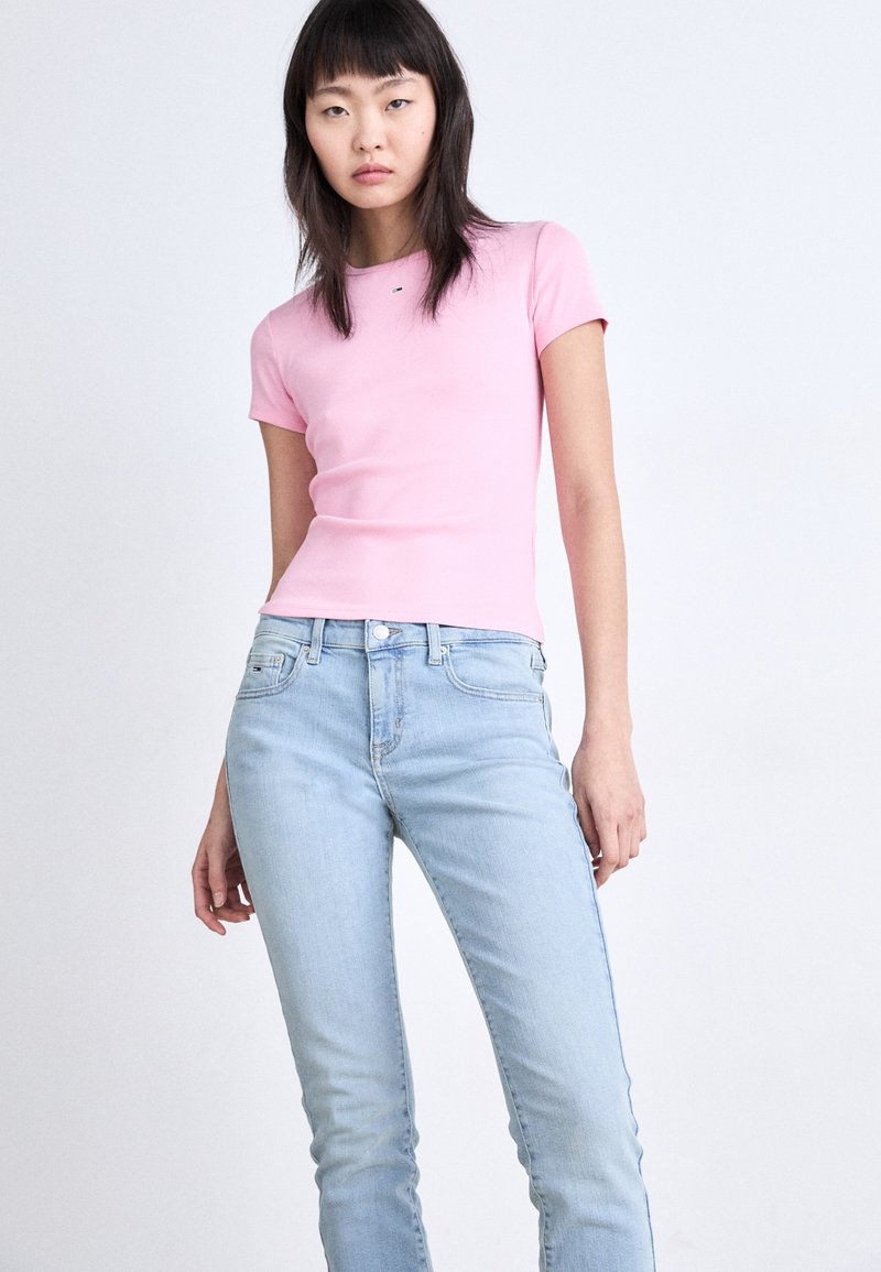 Helles rosa, geripptes T-Shirt mit kurzen Ärmeln, kombiniert mit hellblauen, figurbetonten Jeans. Der Stoff wirkt glatt, mit minimaler Detailverarbeitung.