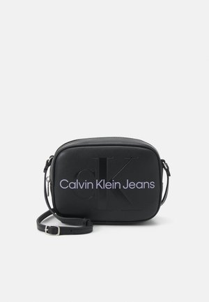 Czarna torebka na ramię Calvin Klein Jeans z wytłoczonym logo "ck" i regulowanym paskiem na jednolitym jasnym tle.