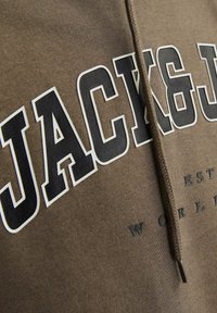 Bruine hoodie met grote zwart-witte tekst "JACK & J" op de voorkant gedrukt. Beschikt over een trekkoord in de capuchon en een gestructureerde stof.