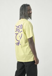 Helles gelbes Baumwoll-T-Shirt mit einem lila abstrakten Design auf der Rückseite. Kurze Ärmel, lockere Passform und runder Ausschnitt.