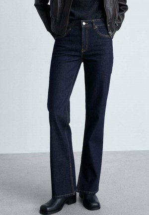 Osoba nosząca granatowe dżinsy o prostych nogawkach, czarne botki na obcasie oraz czarną skórzaną kurtkę z rękami w kieszeniach.