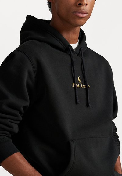 Jauns vīrietis ģērbies melnā Ralph Lauren kapucī, ar zeltītu logotipu un priekšējo kabatu uz vienkārša balta fona.