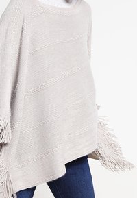Poncho in maglia beige chiaro con accenti di frange, caratterizzato da una vestibilità comoda e strisce testurizzate. Indossato sopra jeans scuri.